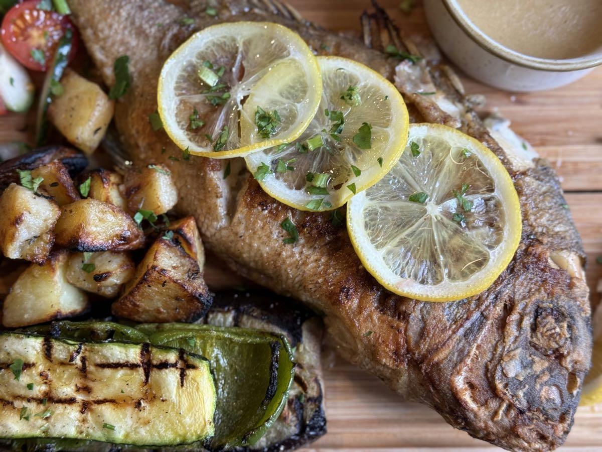 Whole roasted fish with lemon slices and grilled vegetables on a wooden board